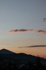 Dramatic mountain silhouette against a serene sunset or sunrise sky, with warm pink and orange clouds and glimpses of a village below.