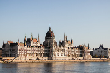 Obraz premium The stunning Hungarian Parliament Building in Budapest, Hungary, stands proudly on the bank of the Danube River, showcasing neo-Gothic architecture against a clear blue sky.