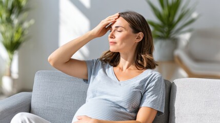A pregnant woman sits on a couch, shielding her eyes from bright sunlight with her hand, feeling warm or relaxed. Her other hand rests on her baby bump.