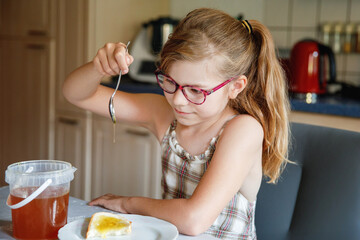 Little school girl with eyeglasses eating bread with honey for breakfast at home. Child enjoying sweet and healthy food in the morning. Real life everyday family lifestyle
