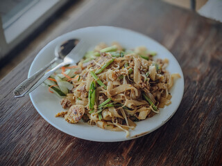 kwetiau noodles with green vegetables on a white plate with a spoon and fork
