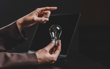 Close-up of hands holding a light bulb in front of a laptop screen, symbolizing innovation and creative concept in a blurred office setting.