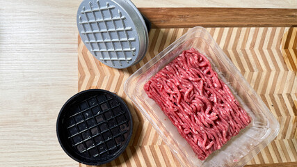 Raw minced beef nestles in a plastic tray beside a burger press, invoking grill flame dreams and Burger Day festivities