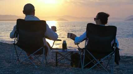 Couple relaxing on a tranquil beach watching the sunset. Relaxed couple sitting in chairs, sipping drinks while watching golden sunlight fading over serene ocean horizon - Powered by Adobe