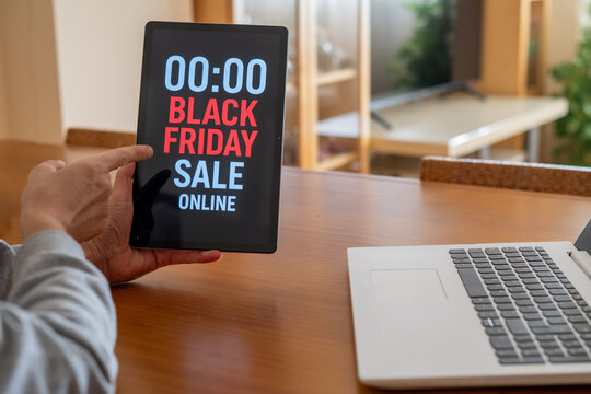 Hands holding tablet displaying black friday online sale countdown at midnight with laptop nearby, suggesting last-minute shopping and e-commerce trends