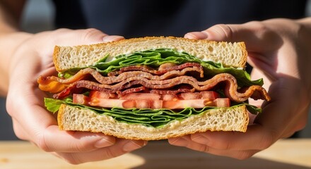 Two hands hold a thick sandwich filled with crispy bacon fresh green lettuce and red tomato slices between toasted bread and creamy spread