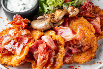 Crispy potato pancakes topped with bacon and sautéed mushrooms, served with creamy herb sauce and fresh greens. Rustic, hearty dish on a restaurant plate