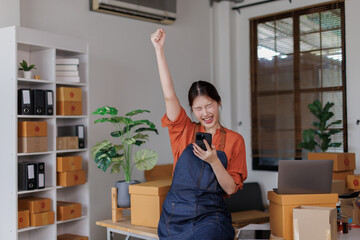 Happy excited asian woman business entrepreneur SME using phone in hand working at home with her laptop,she&rsquo;s online seller SME e-commerce telemarketing concept.
