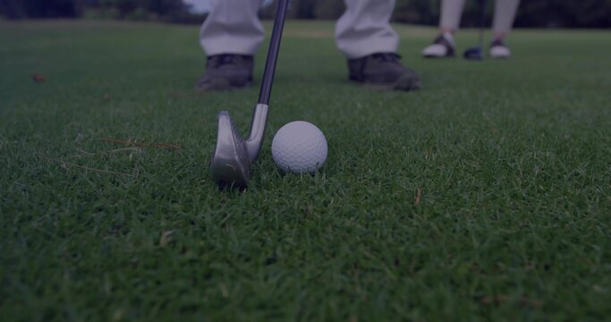 Positioning steel golf iron behind white golf ball on putting green, with golf shoes over grass - Powered by Adobe