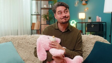 Middle-aged man at home holds pink bunny toy sadly recalling child days feeling tender nostalgia. Young adult guy on sofa misses kid time gentle longing love bittersweet emotion holding plush rabbit