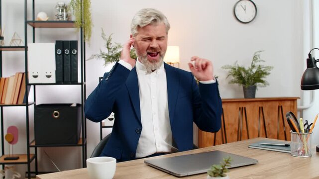 Middle-aged businessman at home office dances on chair wearing earphones singing rock song after working on laptop. Freelancer guy at table writes code enjoying music energy boosting high productivity