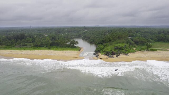 Aerial drone shot flying from over the sea to the golden sands of varkala beach with a backwater inlet and palm coconut trees along coast with monsoon clouds overhead