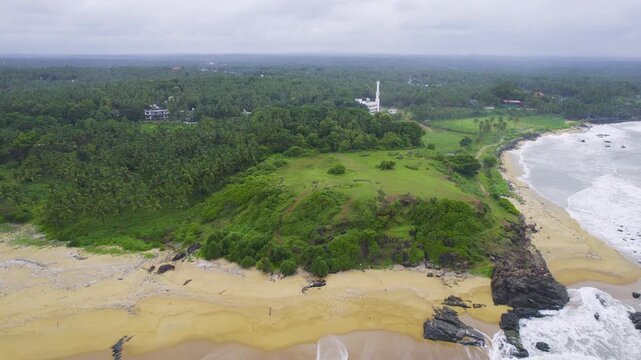 Aerial drone shot flying backwards from a mosque hidden in trees to a wide cliff ledge to the crashing white foam waves at Kappil bekal beach in Kerala