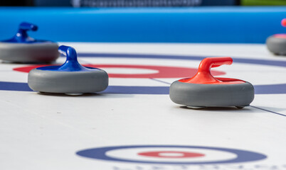 Several curling stones with red and blue handles are positioned on a recreational curling sheet. The setup highlights a friendly game of curling, emphasizing teamwork, strategy, and social engagement