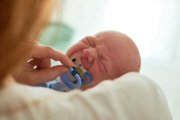 Caucasian infant crying while being comforted by adult woman holding pacifier near mouth, closeup showing newborn facial expression and gentle caregiving interaction