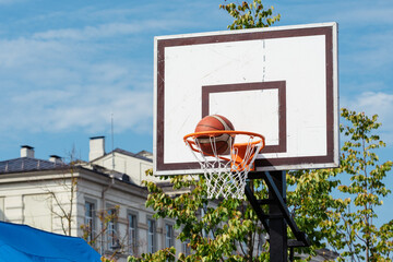 Outdoor basketball hoop with a ball going through the net, set against a clear blue sky and urban buildings in the background © Michele Ursi