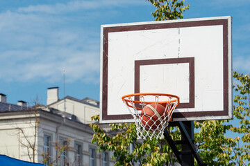Outdoor basketball hoop with a ball going through the net, set against a clear blue sky and urban buildings in the background © Michele Ursi