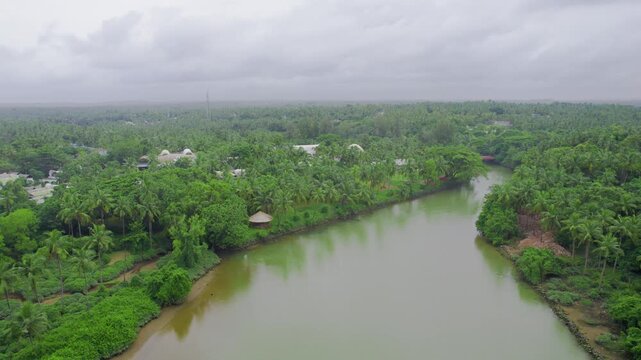 Aerial drone shot flying over backwater canal to buildings in the middle of coconut trees with domed roofs of the Taj hotel under monsoon clouds showing Kappil Bekal Kerala
