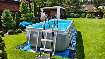 On the mowed grassy lawn stands a gray rectangular PVC frame pool with a ladder. The water in it is filtered. Nearby is a wooden gazebo, swings, bushes and trees. Sunny summer weather and blue sky