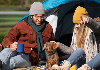 Young couple relaxing in front of tent with their dachshund dog, enjoying hot drinks while camping outdoors