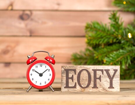 Red alarm clock beside wooden block letters "EOFY" on a rustic wooden surface with Christmas tree branches