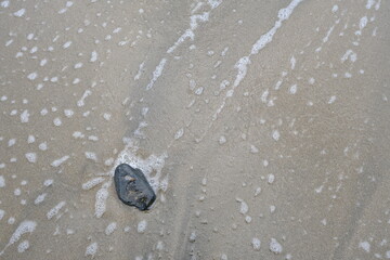 A lone stone rests on smooth sandy beach with ocean waves approaching, symbolizing solitude, calmness, and natural coastal scenery.
