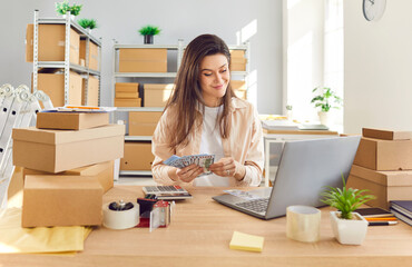 Successful positive woman is seen working at an online store warehouse or post office. She is using a laptop for work and counting cash money, managing storage and profit effectively.