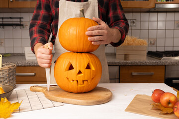 Individual in plaid shirt and apron skillfully carving a stacked pumpkin with a knife on a wooden board, surrounded by autumn leaves and fresh apples, celebrating seasonal festivities