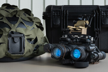 Modern binocular night vision device (NVG) with its mount, displayed next to a camouflaged military helmet and a protective case.
