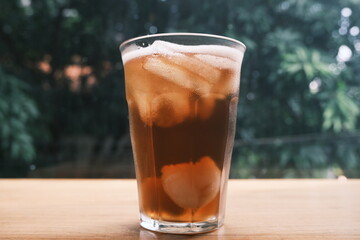Close-up of a glass filled with iced tea and ice cubes placed on a wooden table with greenery in the background.  
