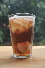Chilled glass of iced tea with lychee fruit and ice cubes, placed on wooden surface with natural green background.
