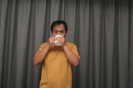 A man in a yellow shirt stands in front of gray curtains, holding and sipping from a white cup, appearing calm and relaxed.  
