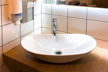 Modern white oval sink on wooden counter in tiled bathroom, chrome faucet. Soap dispenser and paper towel holder mount on wall with mirror. Clean space evokes hygiene and contemporary design.