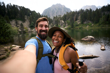 Smiling couple on a summer hike taking a selfie beside a calm mountain lake, wearing backpacks and outdoor gear, enjoying nature, travel adventure, and shared connection in a scenic alpine landscape.