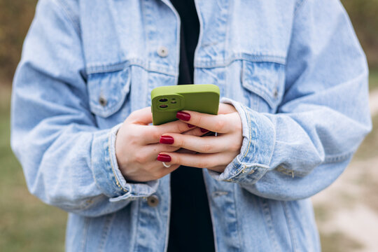 Female hands with red manicure holding green phone in denim jacket.