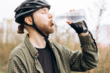 Young bearded man in casual clothes wearing bike helmet drinking water.