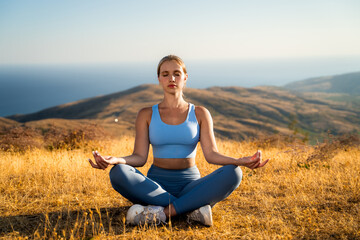 Woman meditating outdoors, front view of peaceful female sitting in lotus yoga pose on mountain meadow at sunset with sea background, wear blue sports, active healthy lifestyle in nature