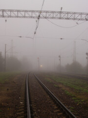 Fototapeta premium Railway tracks disappearing into thick fog with faint train light in distance.