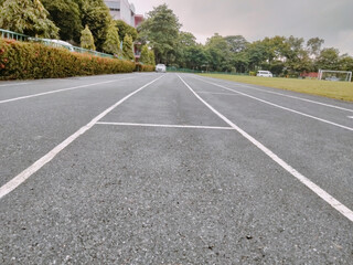 Running track lane lines leading toward distant trees and parked cars under cloudy sky, calm outdoor sports scene