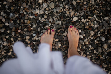 Women's feet with bright pedicure stand on pebbles in shallow waves, wearing a flowing white dress. Scratches on skin, feet slightly wet, sunlight reflecting on water, relaxing beach scene.
