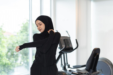 Muslim woman wearing sports hijab stretching arm in gym, showing modern Islamic lifestyle, health, fitness and active living with cultural identity in contemporary environment.