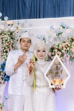 A full-length vertical portrait of an Indonesian Muslim bridal couple standing in all-white traditional attire on their decorated stage, holding the dowry "Mahar" and the official marriage books.