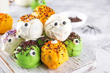 Homemade funny monster mini cakes or cake pops in a colored chocolate glaze and sugar sprinkles. Halloween treat on a concrete background.