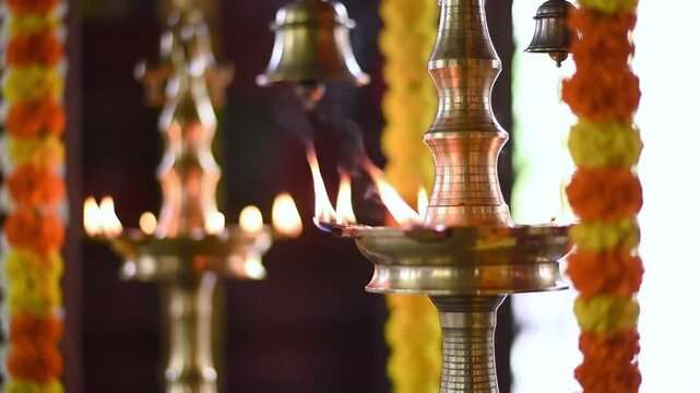 Ornate brass Indian oil lamps with burning flames and hanging bells, decorated with marigold garlands for a ceremony.