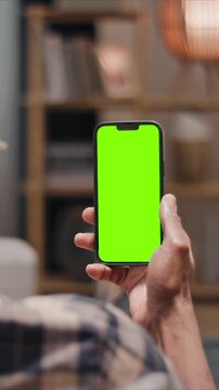 Man holding smartphone with green screen close up, mobile phone in hand, using chroma key mockup indoors at home. Male person browsing blank screen device in vertical position.