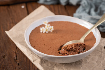 Rich dark chocolate mousse in a white bowl, a spoonful lifted, garnished with delicate white flowers, resting on rustic parchment paper and a wooden surface