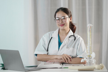 Asian female medical student studying anatomy at desk with textbook, laptop, and spine model, wearing lab coat and glasses. Concept of healthcare education, learning, academic success, and science.