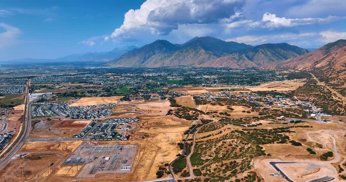 Flight above the deserted land in combination with the urban areas. Cityscape of Spanish Fork, Utah, USA. Fluffy cloudscape in the sky over the rocks.
