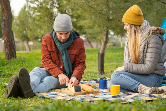 Young couple having a spanish picnic in a park near their tent, the man is cutting cheese on a wooden board