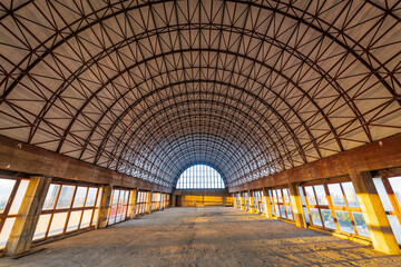 Interior view of an unfinished industrial building with a large arched steel-truss roof structure and expansive floor-to-ceiling windows. 